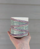 Alternate side view of 3D printed porcelain ceramic cup. Held in the palm of a hand. Cup has 4 textured rings draped diagonally around the cup. Rings are glazed translucent lavender. The top of the cup is glazed white. The background of the cup is glazed translucent sage green. 