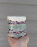 Side View of 3D printed porcelain ceramic cup. Held in the palm of a hand. Cup has 4 textured rings draped diagonally around the cup crossing at 2 points. Rings are glazed translucent lavender. The top of the cup is glazed white. The background of the cup is glazed translucent sage green.