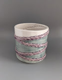Slightly angled from the top view of 3D printed porcelain ceramic cup. Held in the palm of a hand. Cup has 4 textured rings draped diagonally around the cup. Rings are glazed translucent lavender. The top of the cup is glazed white. The background of the cup is glazed translucent sage green.
