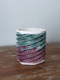 Close up side view of 3D printed porcelain cup. on a wood table It is textured with tight spirals all the way up. The bottom half is glazed a translucent dark berry, the top half is glazed a translucent teal. The top ring is glazed white.