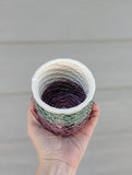 Angled top down view in the palm of a hand of 3D printed porcelain cup. It is textured with tight spirals all the way up. The bottom half is glazed a translucent dark berry, the top half is glazed a translucent teal. The top ring is glazed white. The inside is glazed white in the top half and translucent dark berry the bottom half.